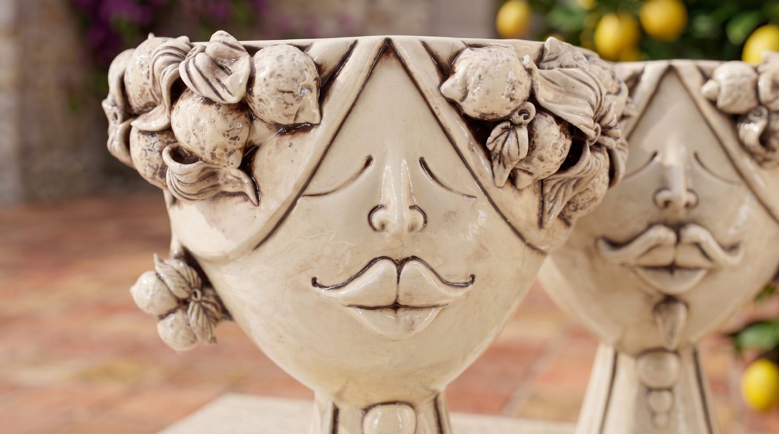 Close-up of two cream-white ceramic heads with modeled lemons in their crowns, in front of a lemon tree