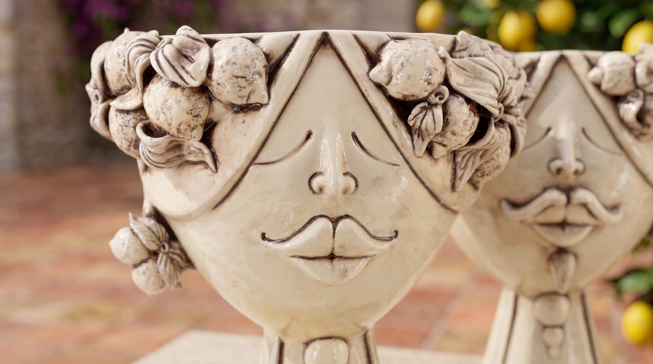 Close-up of two cream-white ceramic heads with modeled lemons in their crowns, in front of a lemon tree